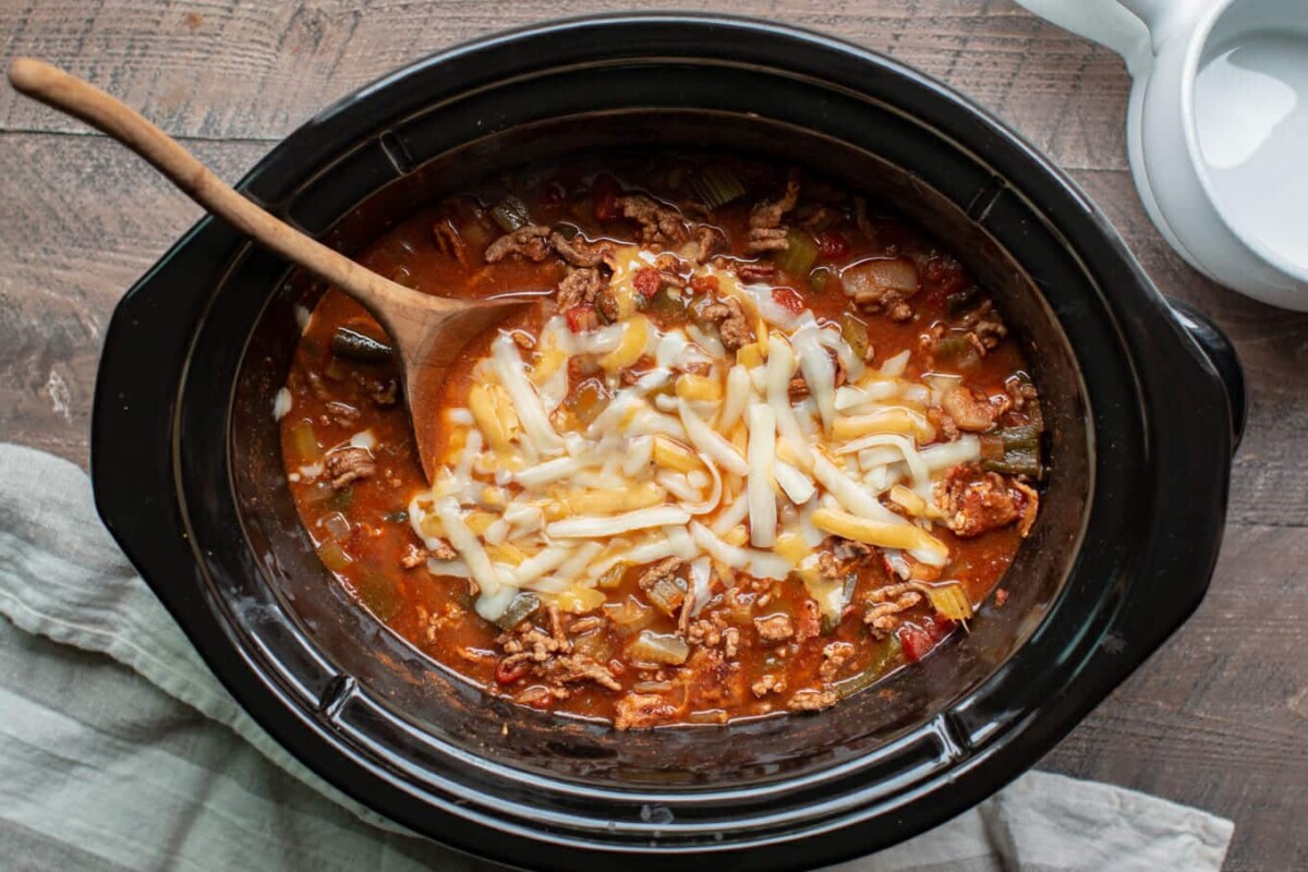 A top view of a slow cooker filled with chili topped with melted cheese. A wooden spoon rests inside, with a beige cloth nearby. The chili contains visible chunks of vegetables and meat in a reddish broth.
