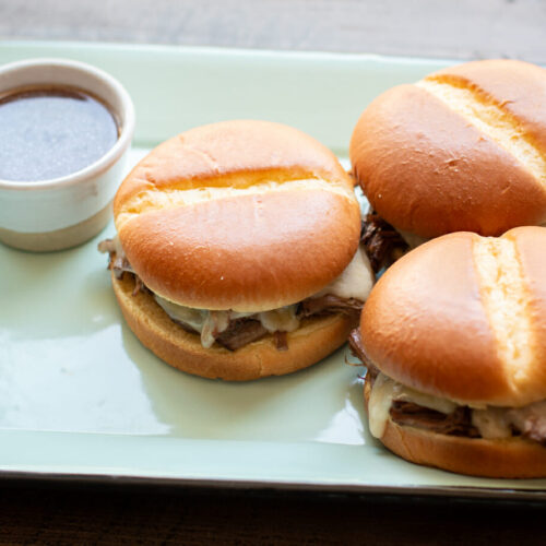 3 french dip sandwiches on a teal tray with au jus on the side.