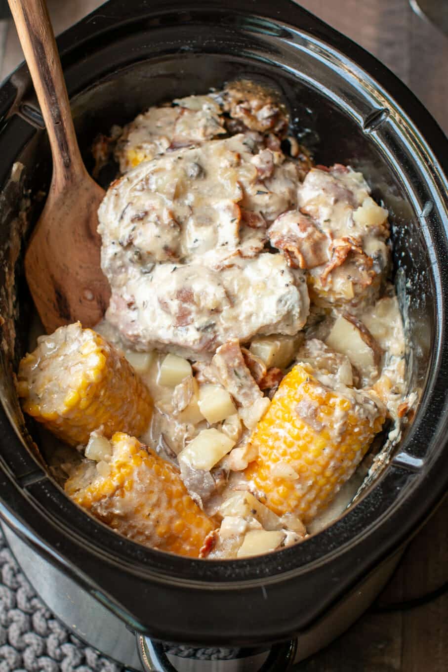 cooked pork chops, corn cobs, potatoes with spatula in slow cooker.