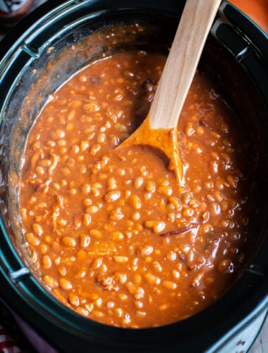 close up of root beer baked beans with wooden ladle in slow cooker