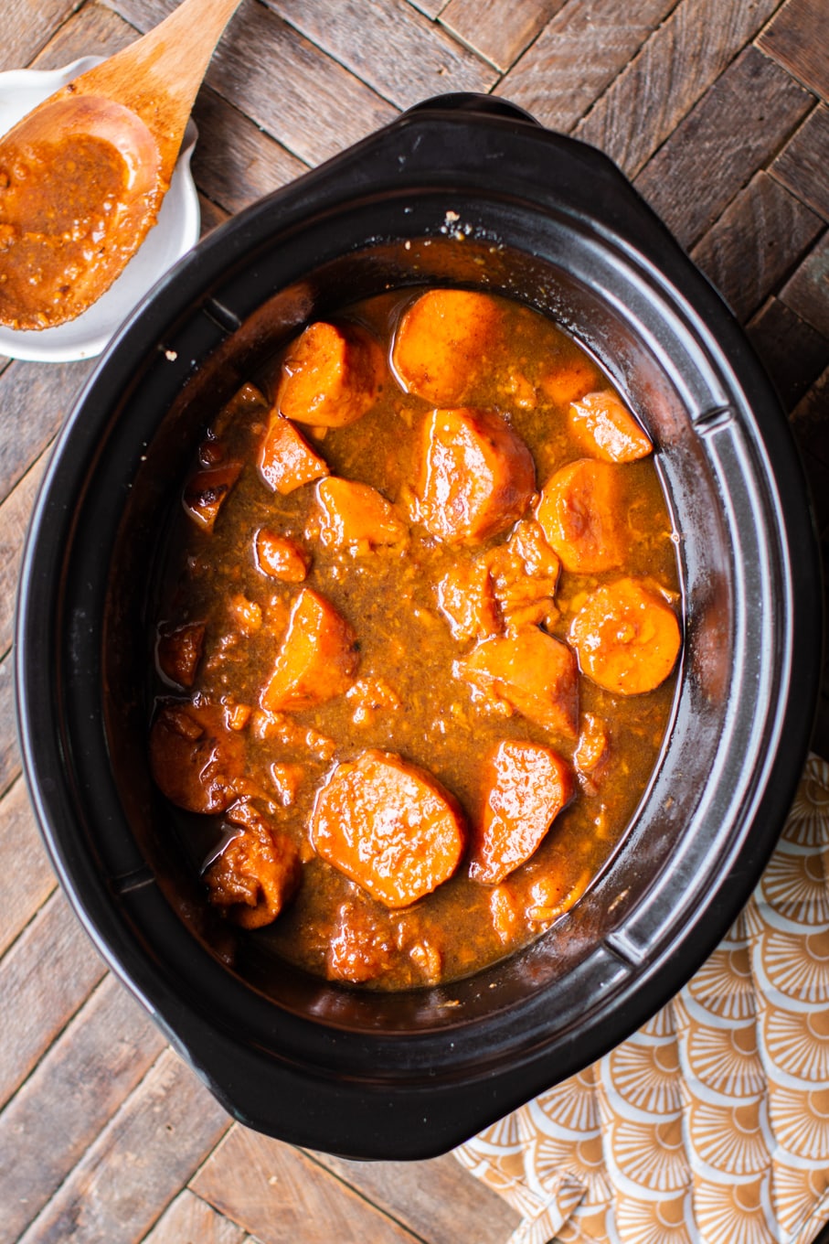 Candied yams in the slow cooker in rich brown sauce with orange pot holder next to it.