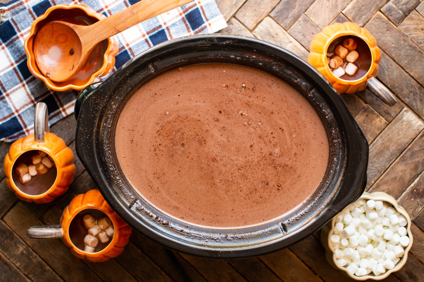 rich and creamy hot chocolate in slow cooker with pumpkin mugs around it.