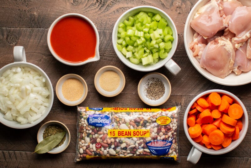 onion, celery, carrots, a bag of 15 bean soup mix, seasonings and chicken thighs on a wooden table.