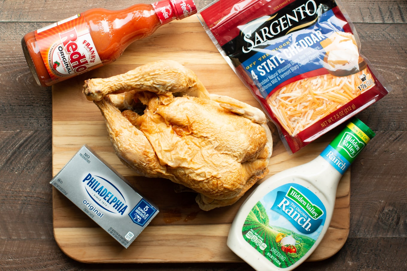 rotisserie chicken, cheese, ranch dressing, cream cheese, and buffalo sauce on a cutting board.