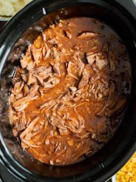 Shredded meat in sauce in a slwo cooker with mashed potatoes and corn on the side.