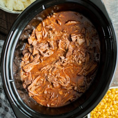 Shredded meat in sauce in a slwo cooker with mashed potatoes and corn on the side.