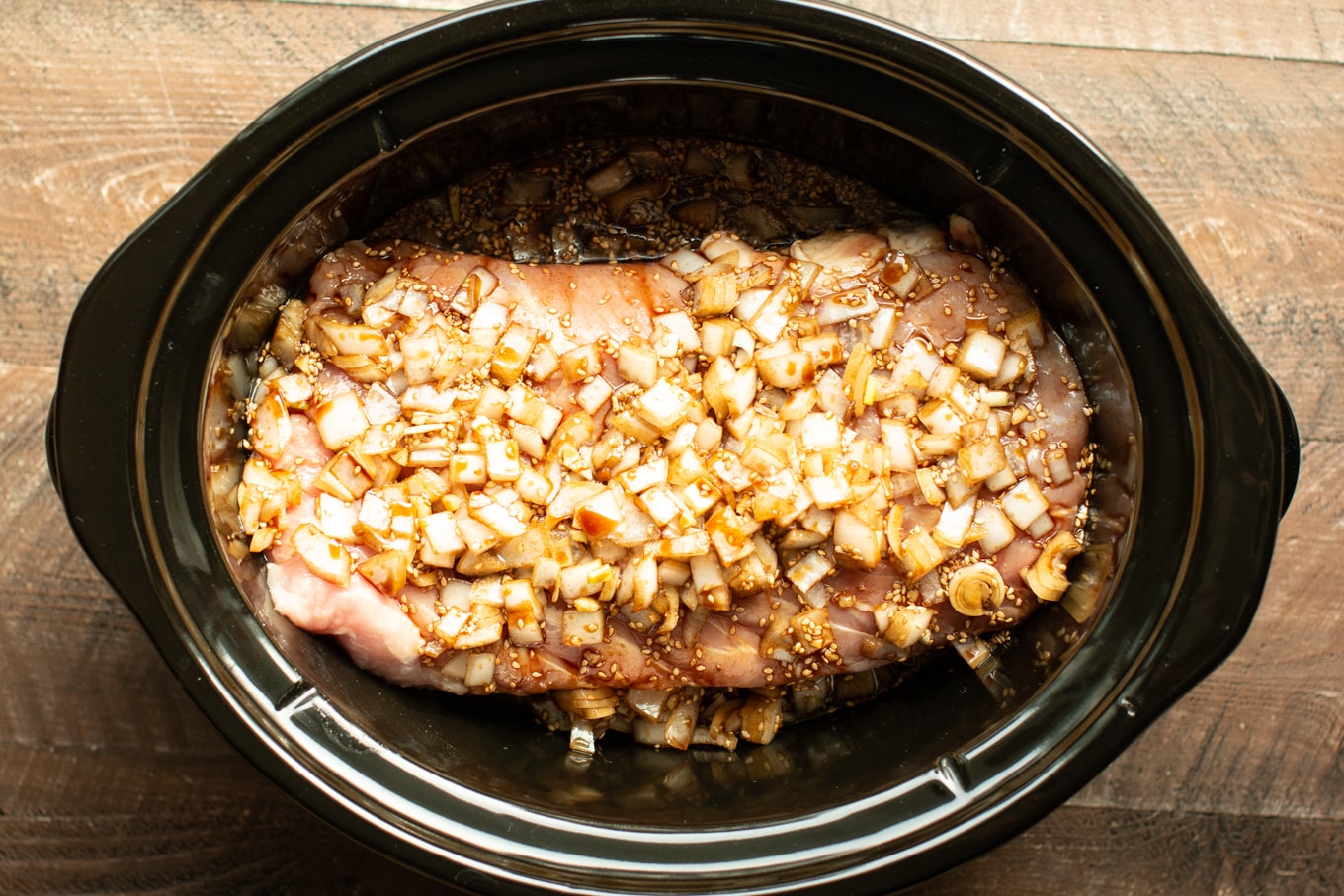 uncooked pork loin in a slow cooker with onions, soy, brown sugar and sesame seeds.