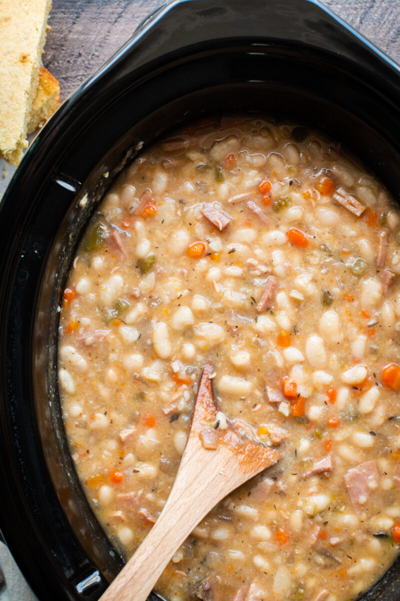 close up photo of white beans and ham with wooden spoon in them.