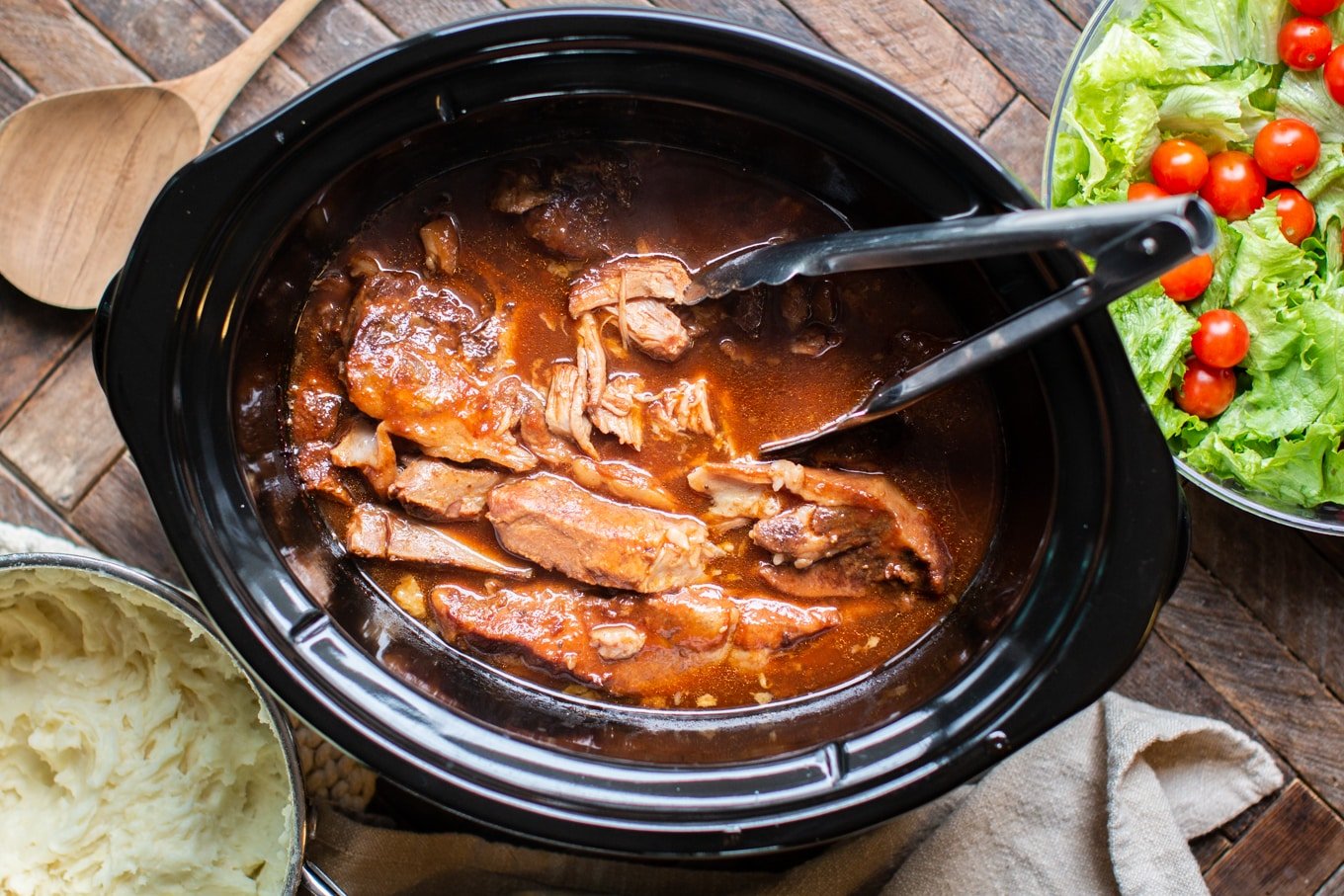 pork ribs in sauce in slow cooker with tongs in it.