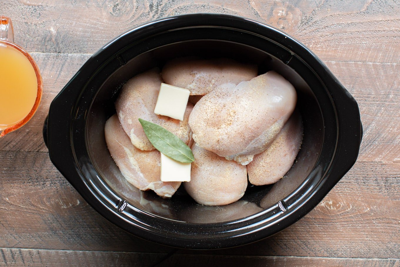 raw chicken breasts in an oval slow cooker with butter pats, bay leaf and pepper on top.