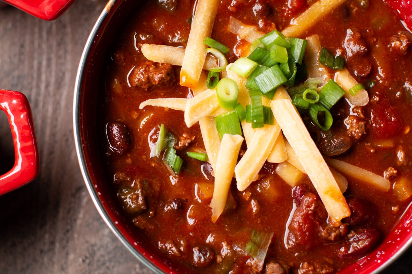 Close up of venison chili in a red bowl with cheddar cheese and green onion on top.