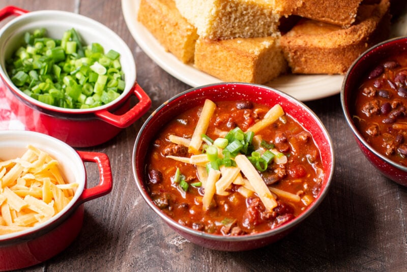 Red bowl of chili with cheese and onions, cornbread in the background.