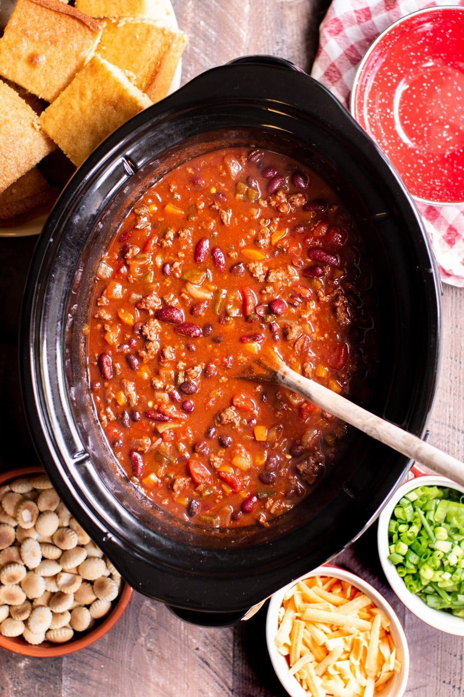 Venison chili in a slow cooker with cornbread, oyster crackers, green onion and cheese on the side.