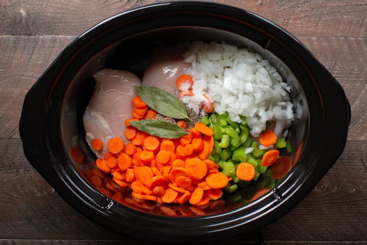 Uncooked chicken, sliced carrots, celery, diced onions, bay leaves and pepper in a slow cooker.