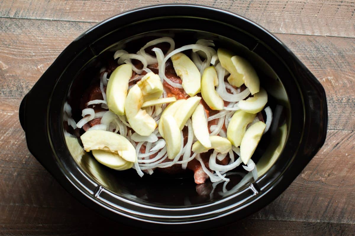 apples and onions on top of apple butter and pork chops in the slow cooker, uncooked.