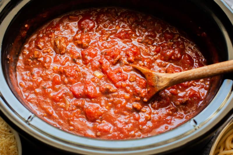 close up of cooked meaty spaghetti sauce in a slow cooker with a wooden spoon in it.