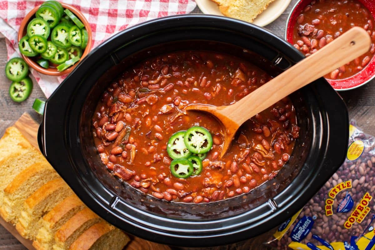 serving cooked barbecue beans with a wooden spoon, jalapenos and cornbread on the side.