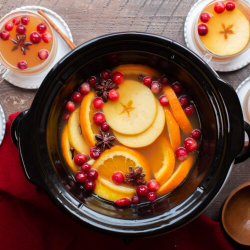 Finished apple cider in slow cooker with cider in mugs around the sides.