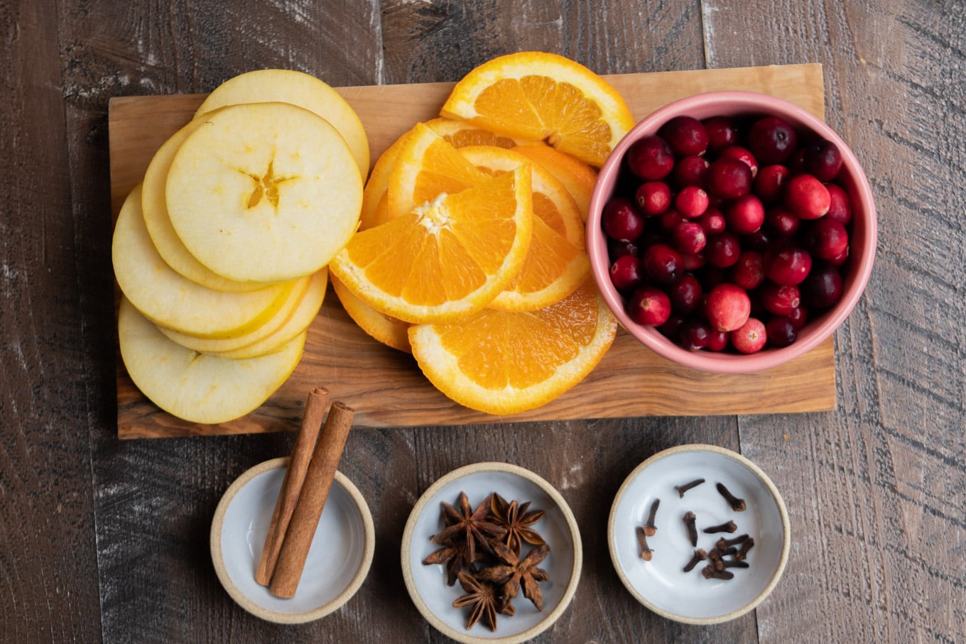 slice apples, sliced oranges, pink bowl of cranberries. Spices in small white dishes.