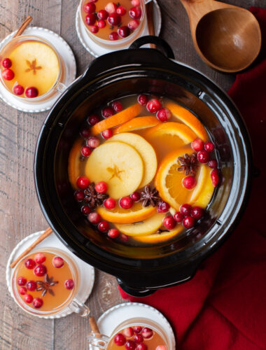 apple cider in round slow cooker with 4 clear cups of cider around the side.