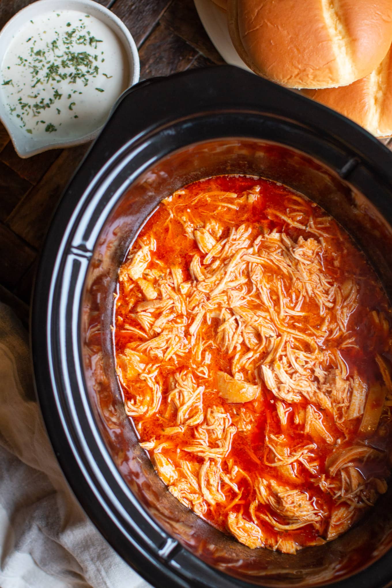 shredded buffalo chicken in slow cooker with ranch and buns on the side.