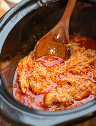 close up of chicken in buffalo sauce in the slow cooker, with wooden spoon dipping in.