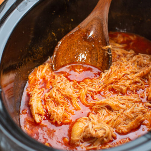 close up of chicken in buffalo sauce in the slow cooker, with wooden spoon dipping in.