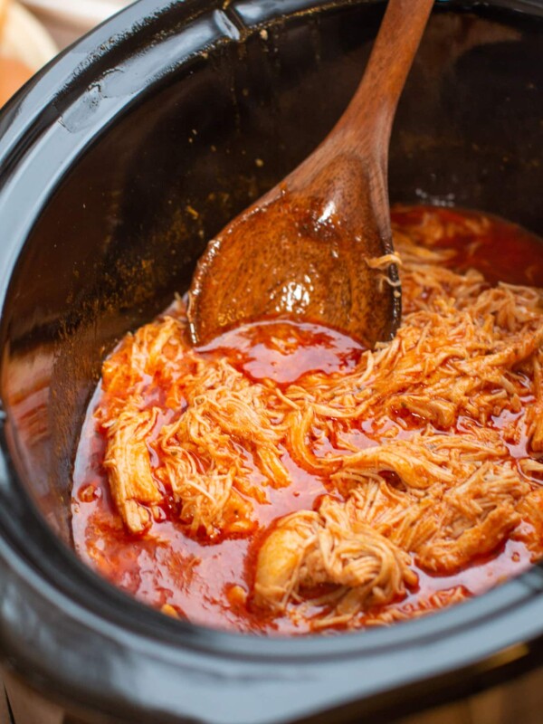 close up of chicken in buffalo sauce in the slow cooker, with wooden spoon dipping in.