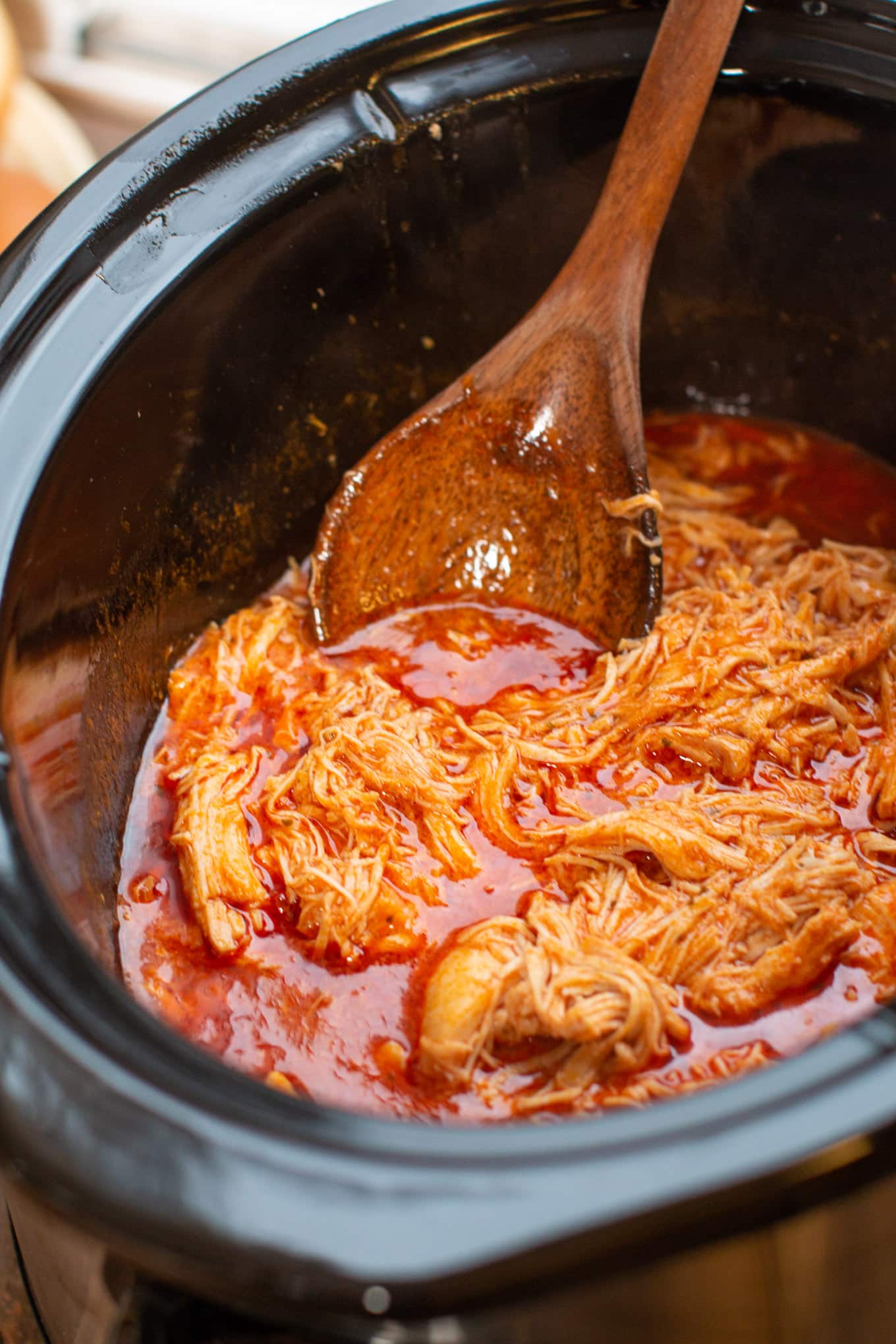 close up of chicken in buffalo sauce in the slow cooker, with wooden spoon dipping in.
