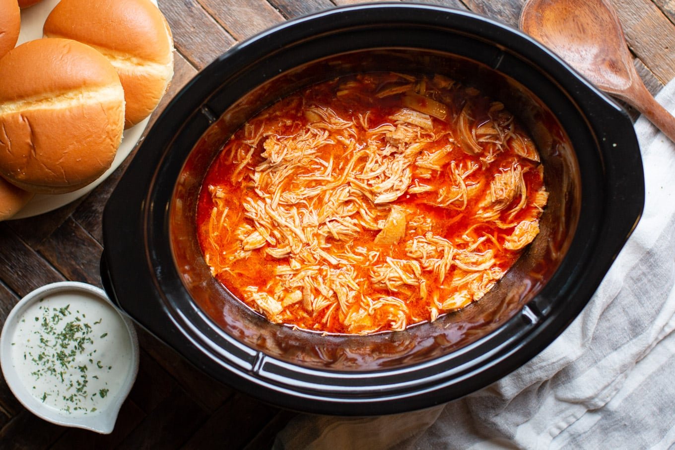 shredded chicken in slow cooker with buns, ranch, and wooden spoon on side.