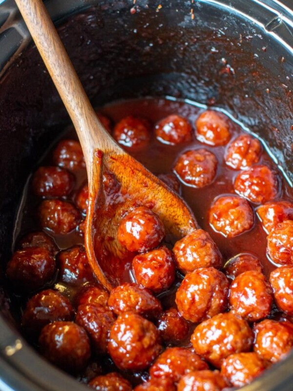 Grape jelly meatballs in the slow cooker with a wooden spoon.