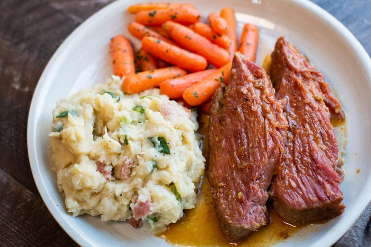 A white plate with mashed potatoes mixed with green herbs and bits of bacon, two slices of braised beef in a brown sauce, and a serving of glazed baby carrots. The dish is arranged neatly, with the vegetables on one side and the meat on the other.