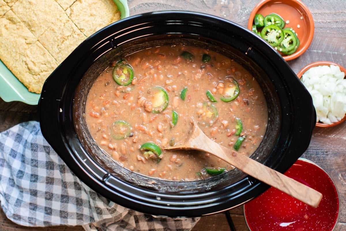 Cooked pinto beans in slow cooker with jalapenos and wooden spoon in them.