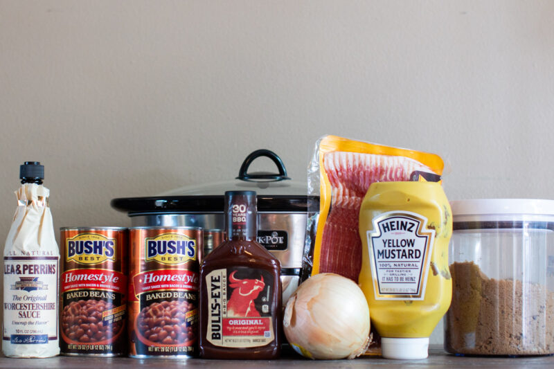 worcestershire sauce, baked beans, barbeucue sauce, onion, bacon, mustard and brown sugar in front of slow cooker.