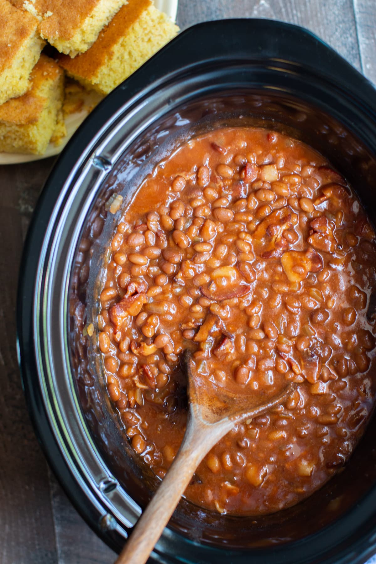baked beans with bacon in black slow cooker with wooden spoon in them.