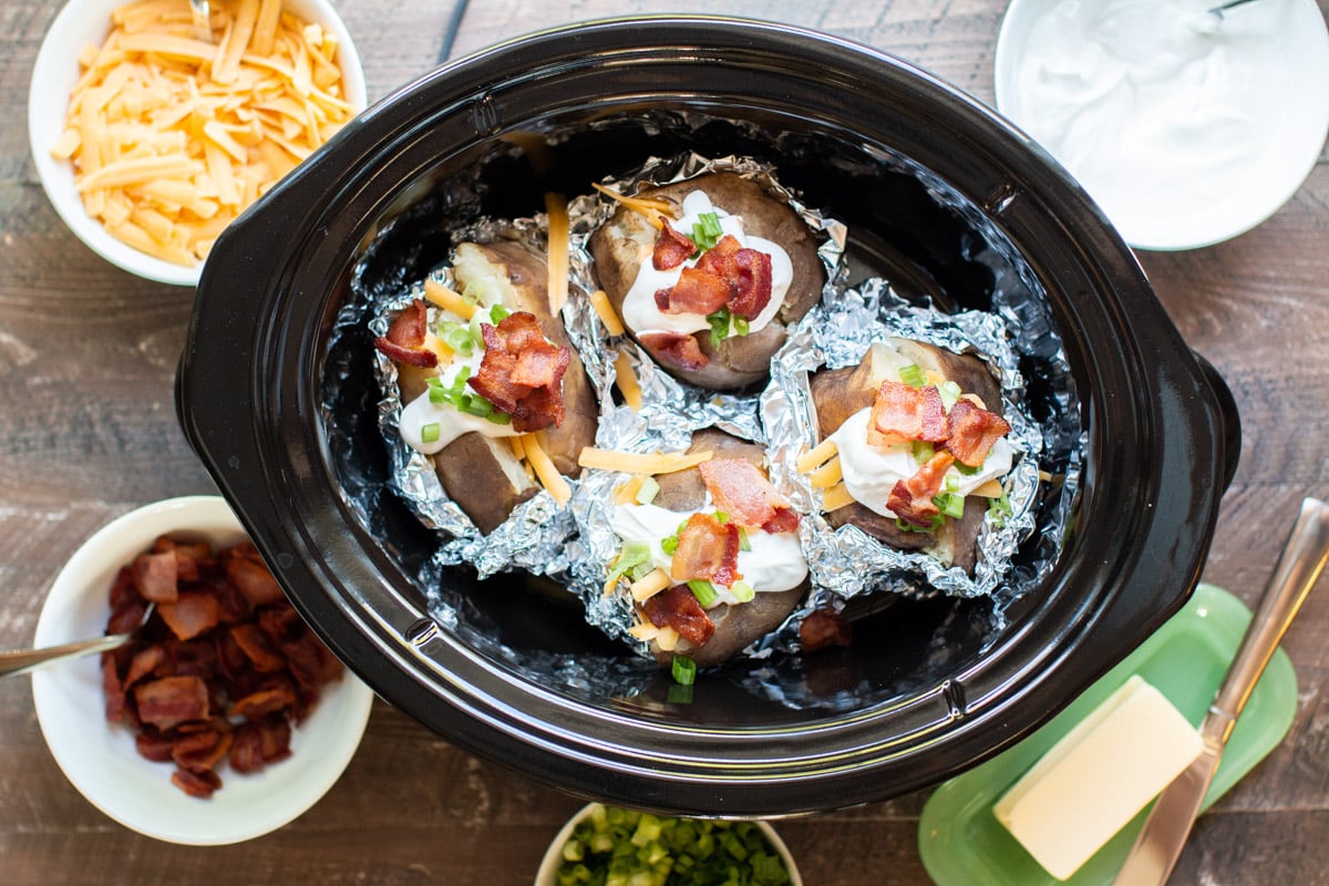 4 baked potatoes in slow cooker halfway unwrapped with toppings on each one