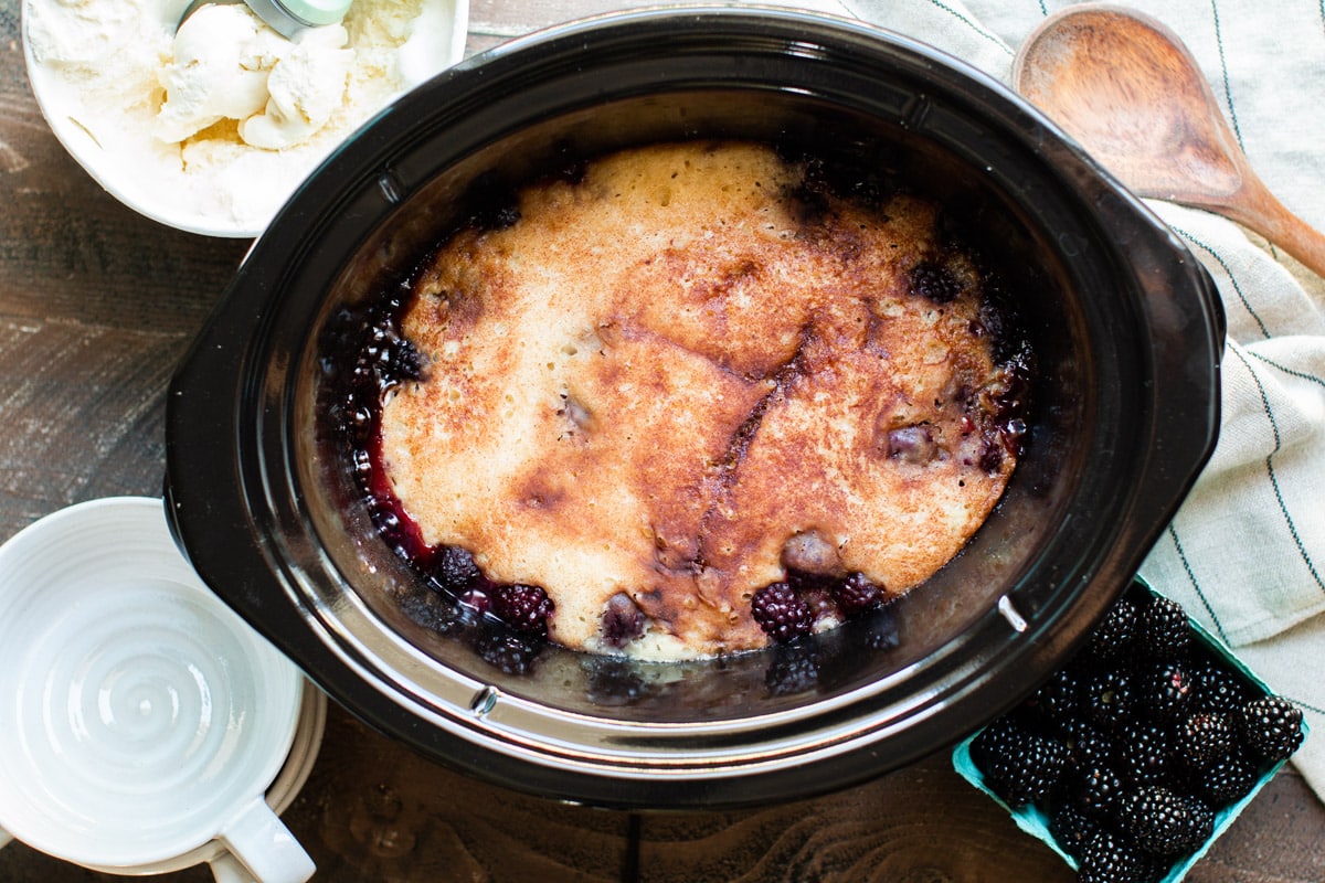 finished cooking blackberry cobbler in the slow cooker. Ice cream, bowls and blackberries on the side.