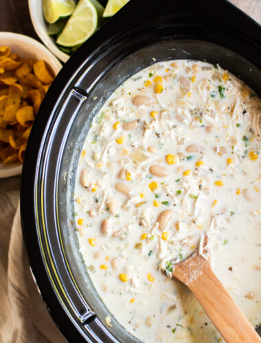 close up of white chicken chili in slow cooker with wooden spoon in it. Fritos on side.