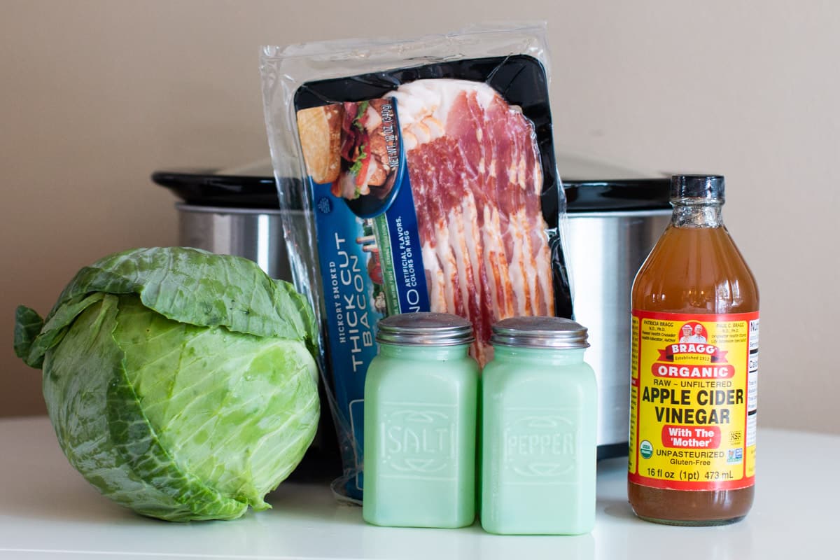 large head cabbage, bacon, salt, pepper, and bacon in front of a slow cooker.