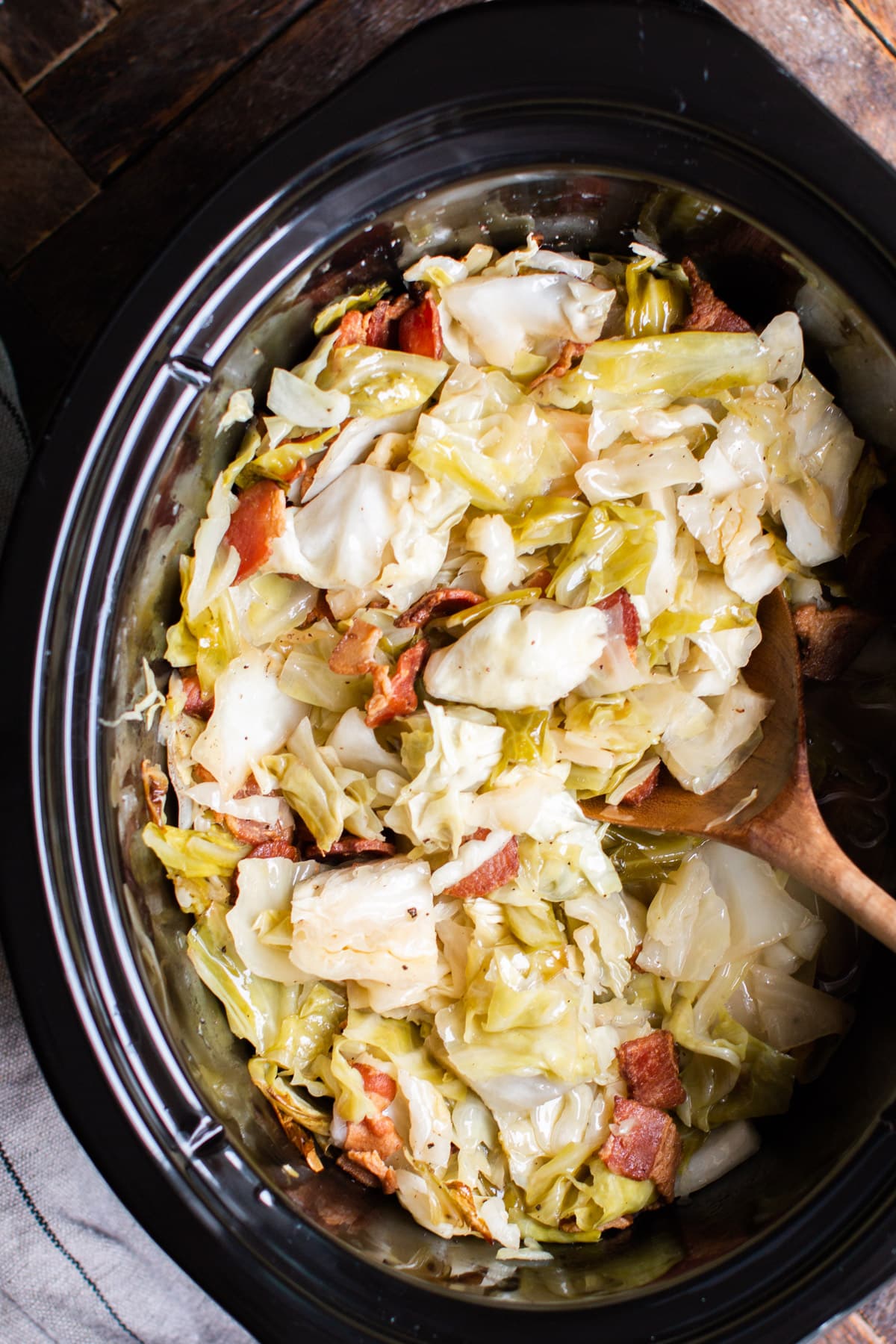 cooked cabbage, bacon in a slow cooker with a wooden spoon in it.