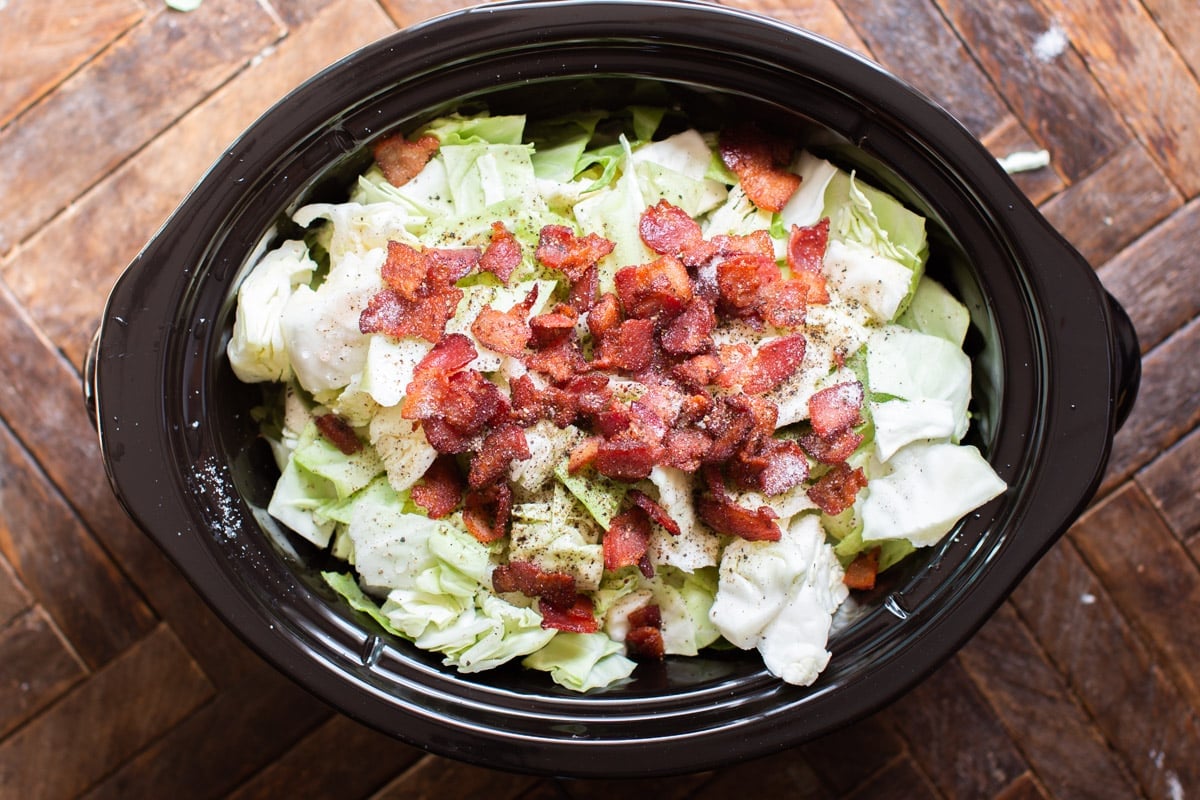 uncooked cabbage in a slow cooker with bacon and pepper on top.