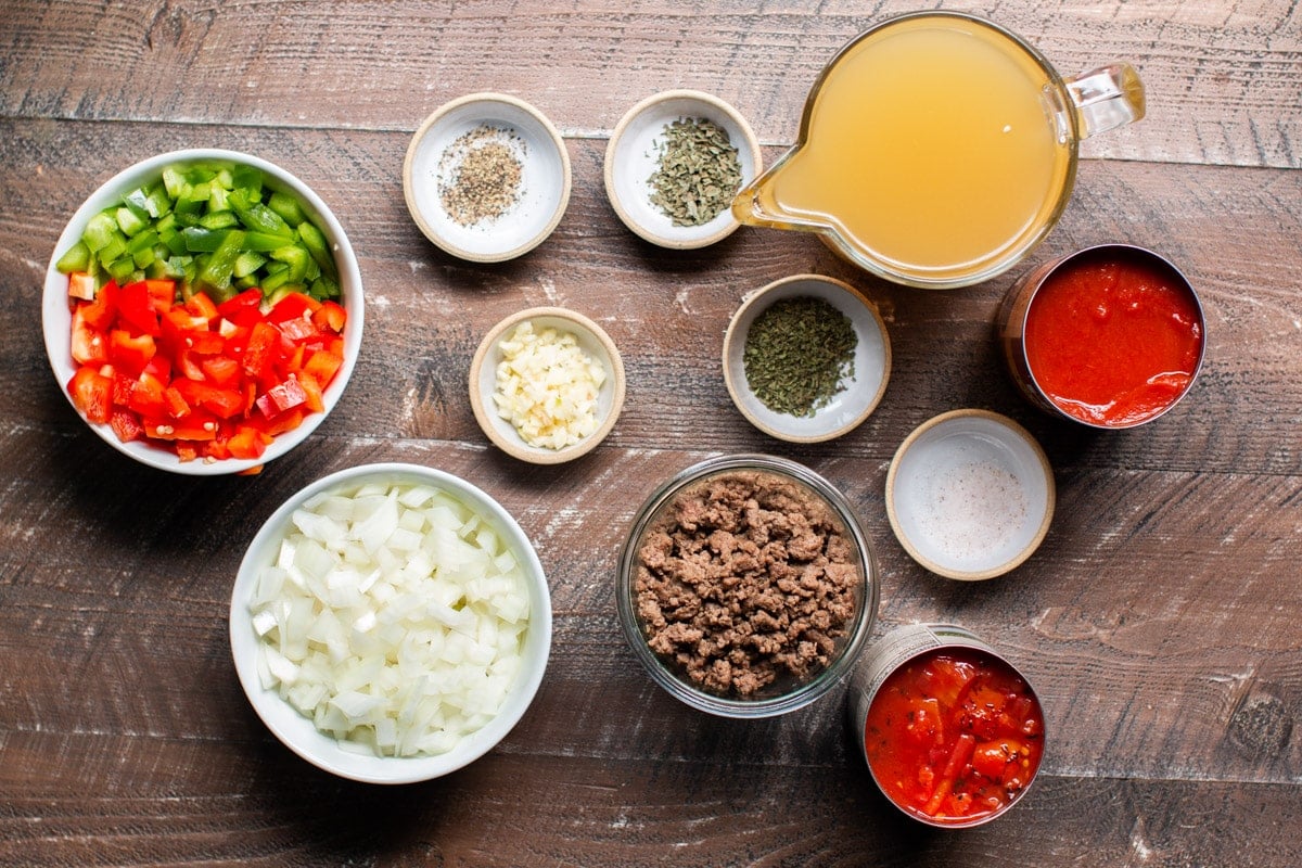 diced bell peppers, onion, garlic, seasonings, broth, ground beef, cans of tomatoes on wooden table.