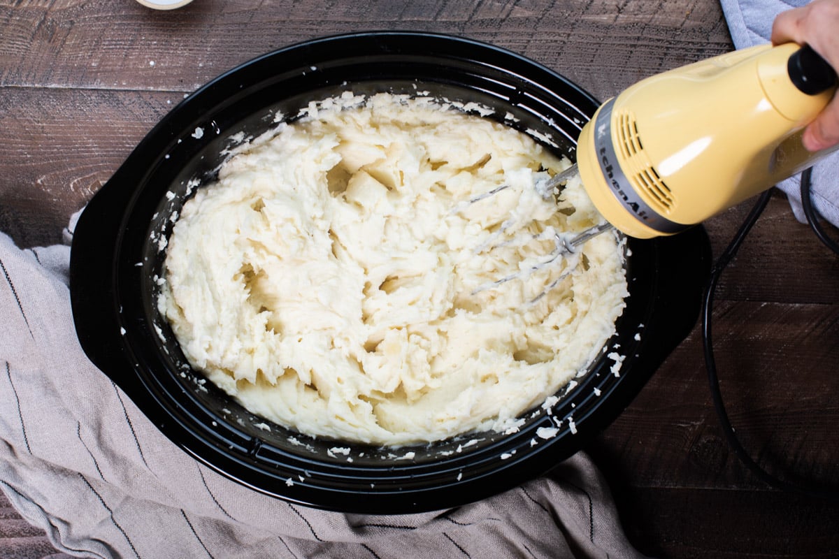 potatoes being blending with yellow hand mixer in slow cooker