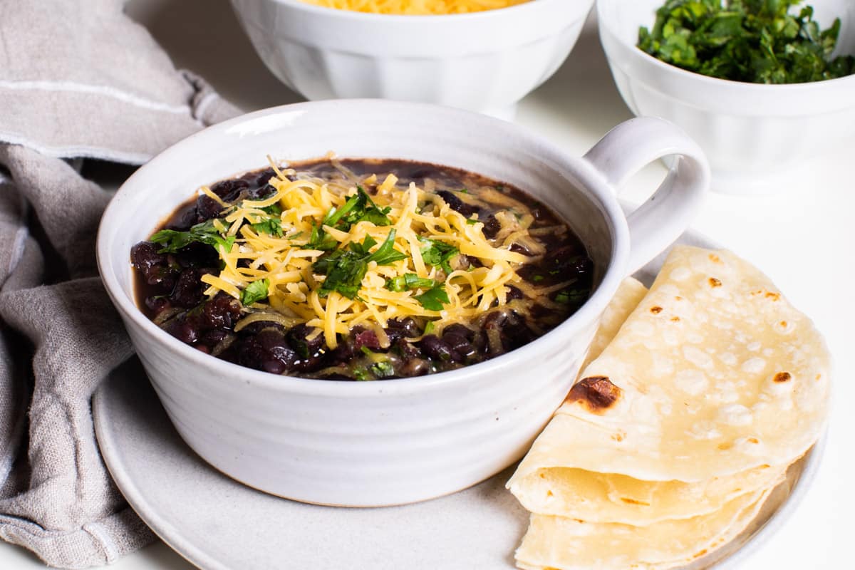 white bowl with black bean soup in it. Topped with cheddar cheese and cilantro.