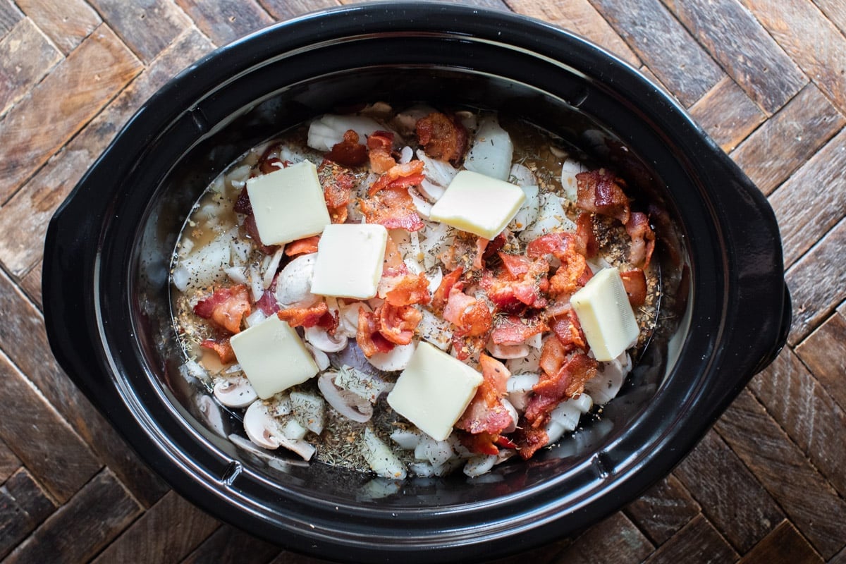 venison roast in slow cooker with bacon, onion, butter and onion on top, before cooking.