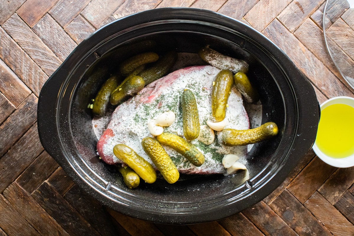 raw chuck roast with seasonings and pickles on top.