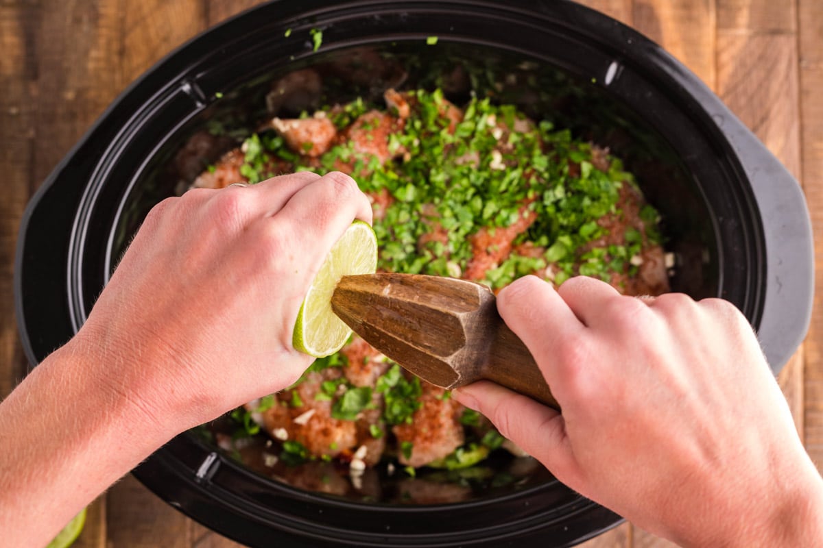 lime juice being squeezed over chicken, peppers, onions and cilantro in slow cooker.