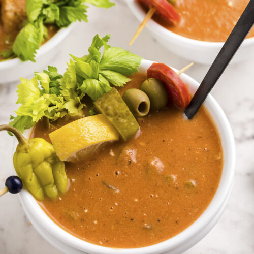 close up of bloody mary soup in bowl with pepper, lemon, pickle, olive and pepperoni on top