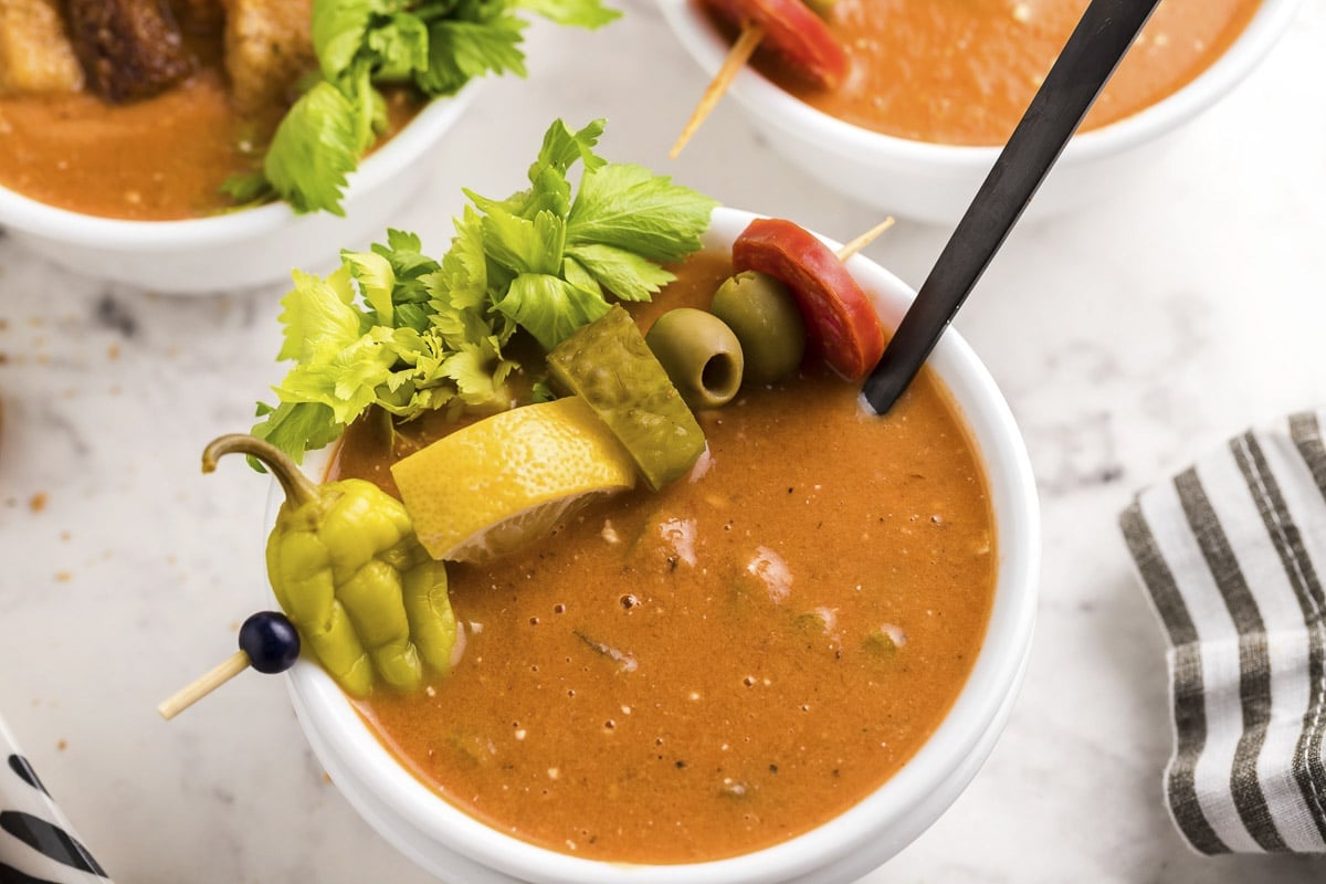 close up of bloody mary soup in bowl with pepper, lemon, pickle, olive and pepperoni on top