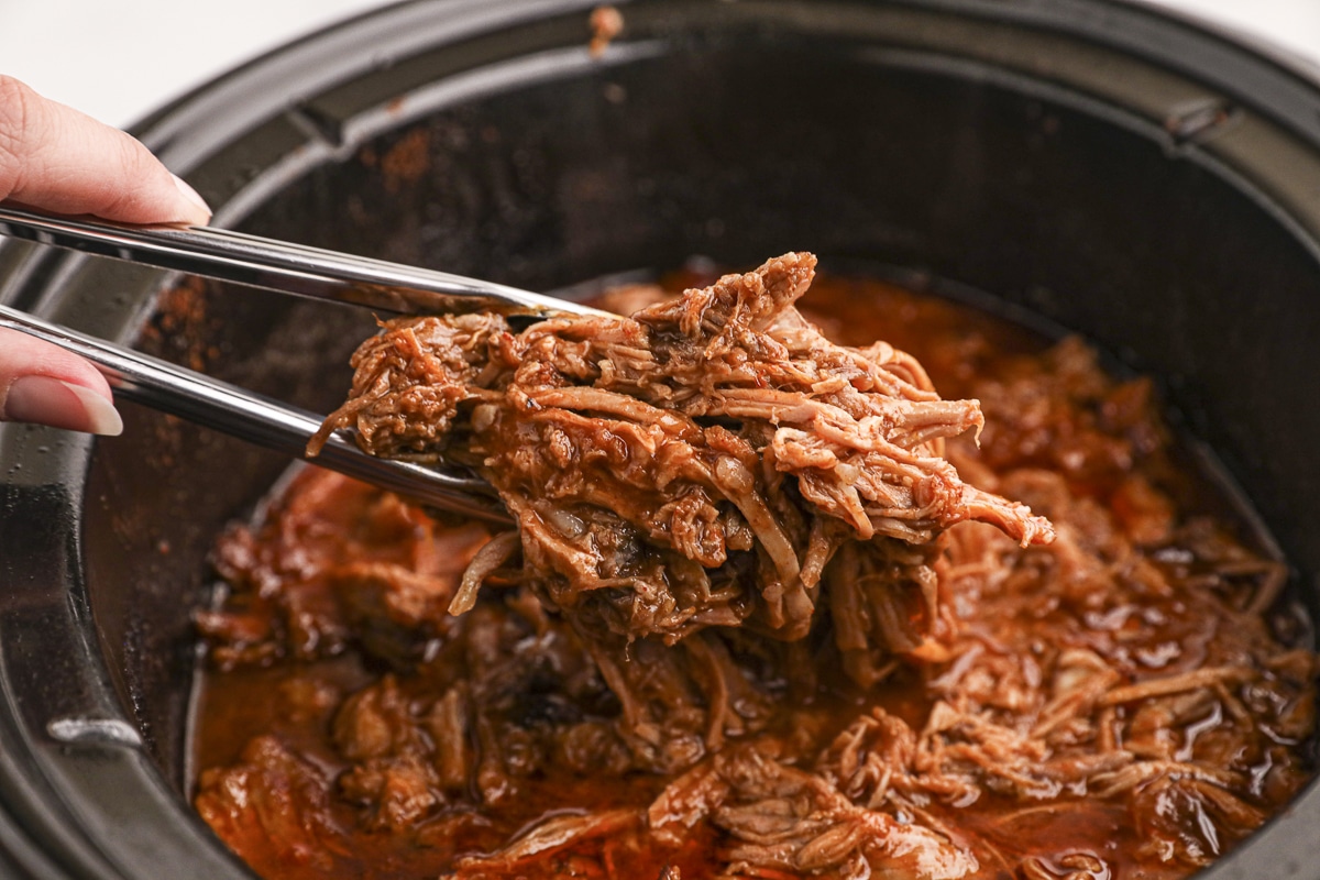 beer pulled pork on tongs coming from slow cooker.